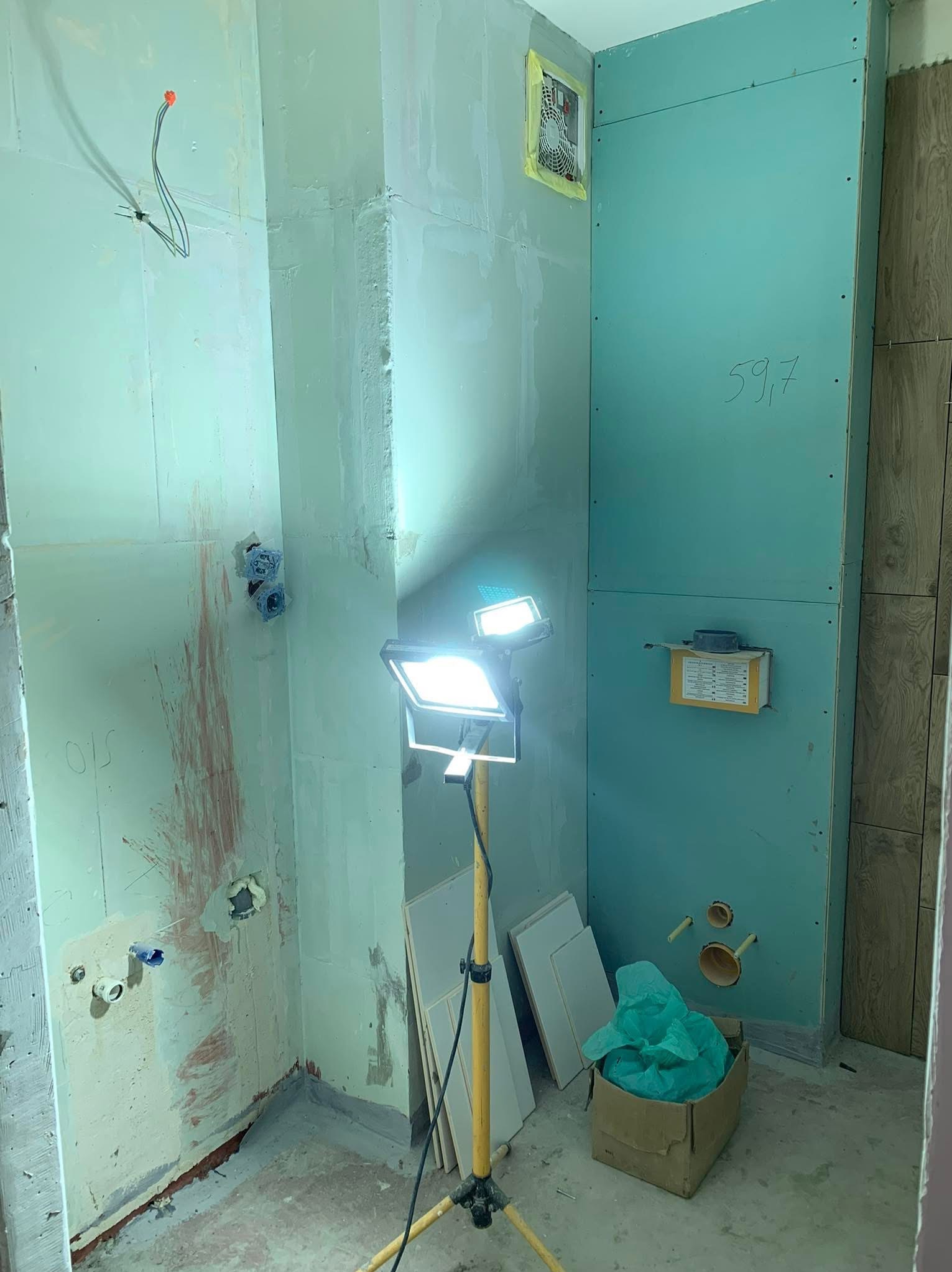 Wnętrze łazienki w trakcie remontu. Widoczne ściany pokryte płytami gipsowymi, instalacje, lampa budowlana i materiały. Na ścianie napis '59,7'.
