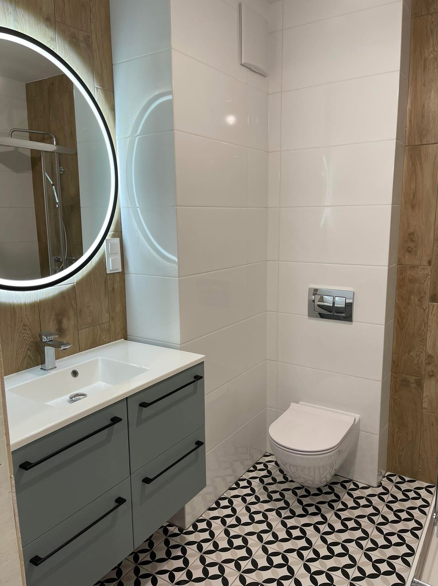 Nowoczesna łazienka z okrągłym lustrem LED, szarą szafką pod umywalką i białą toaletą. Na podłodze wzorzyste, czarno-białe płytki. Ściany wyłożone białymi i drewnopodobnymi płytkami.