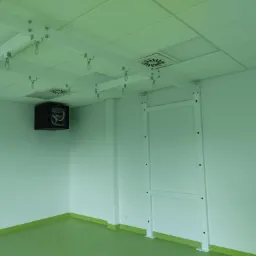 Biała, metalowa konstrukcja sufitowa z hakami w czystym pomieszczeniu o zielonej podłodze, widoczna szafa rack na ścianie.