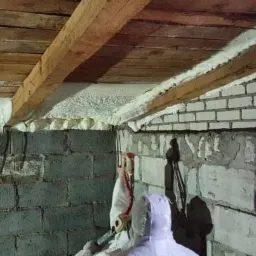 Poddasze w trakcie ocieplania pianką PUR, widoczne drewniane belki stropowe i biała pianka izolacyjna na ścianach z pustaków, pracownik w kombinezonie ochronnym aplikuje pianę.