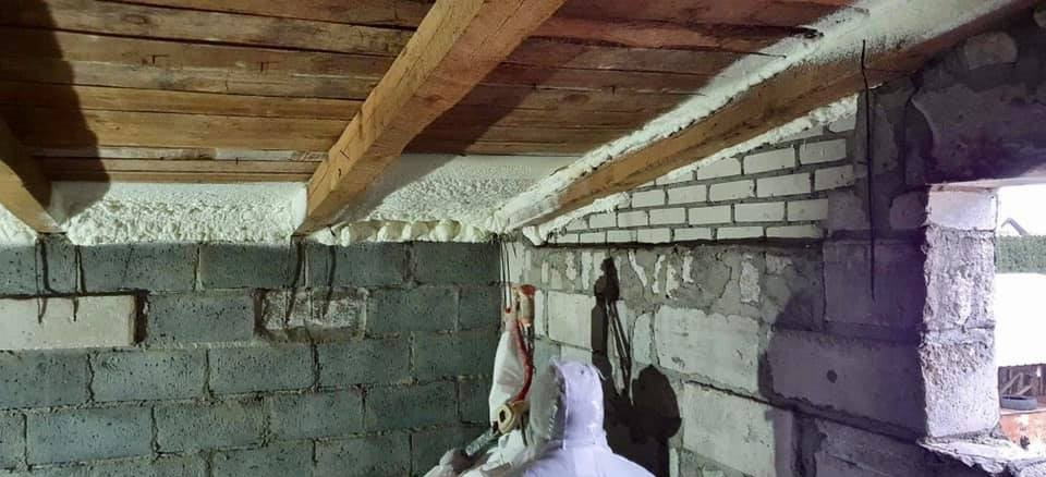 Poddasze w trakcie ocieplania pianką PUR, widoczne drewniane belki stropowe i biała pianka izolacyjna na ścianach z pustaków, pracownik w kombinezonie ochronnym aplikuje pianę.