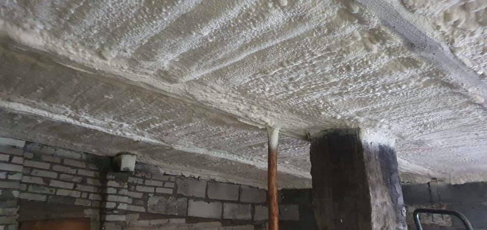 Poddasze z widoczną konstrukcją stropu pokrytą grubą warstwą szarej pianki izolacyjnej, z widocznym słupem konstrukcyjnym i fragmentem ściany z cegły dziurawki.