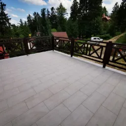 Jasne, prostokątne płytki ułożone na balkonie z drewnianą balustradą na tle lasu i zabudowań.