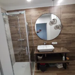 Nowoczesna łazienka z kabiną prysznicową, umywalką nablatową na drewnianym blacie i okrągłym lustrem z wiszącymi lampami; w lustrze odbija się osoba robiąca zdjęcie.