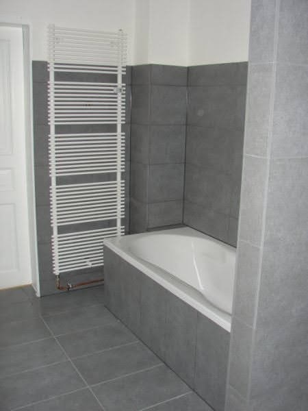 Szara łazienka z białą wanną obudowaną szarymi płytkami, grzejnikiem drabinkowym i szarą podłogą z płytek.