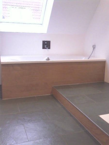 Wykończona łazienka na poddaszu z obudowaną wanną w drewnopodobnym panelu i szarą, kamienną podłogą z niewielkim podestem oraz oknem dachowym.