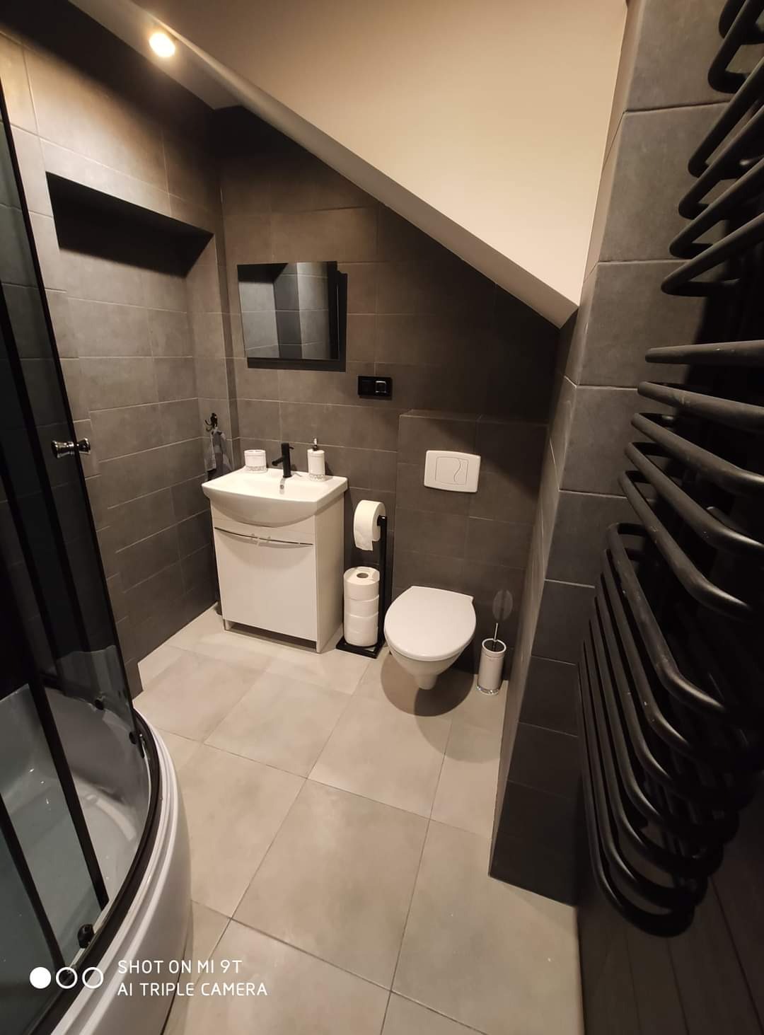 Nowoczesna łazienka w odcieniach szarości z białą armaturą, kabiną prysznicową z czarnymi ramami i grzejnikiem drabinkowym w kolorze czarnym, z ukośnym sufitem.