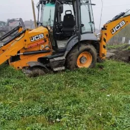 Żółto-szara koparko-ładowarka JCB w trakcie prac ziemnych na trawiastym terenie, widoczne ślady błota na oponach i łyżce, w tle zabudowania.