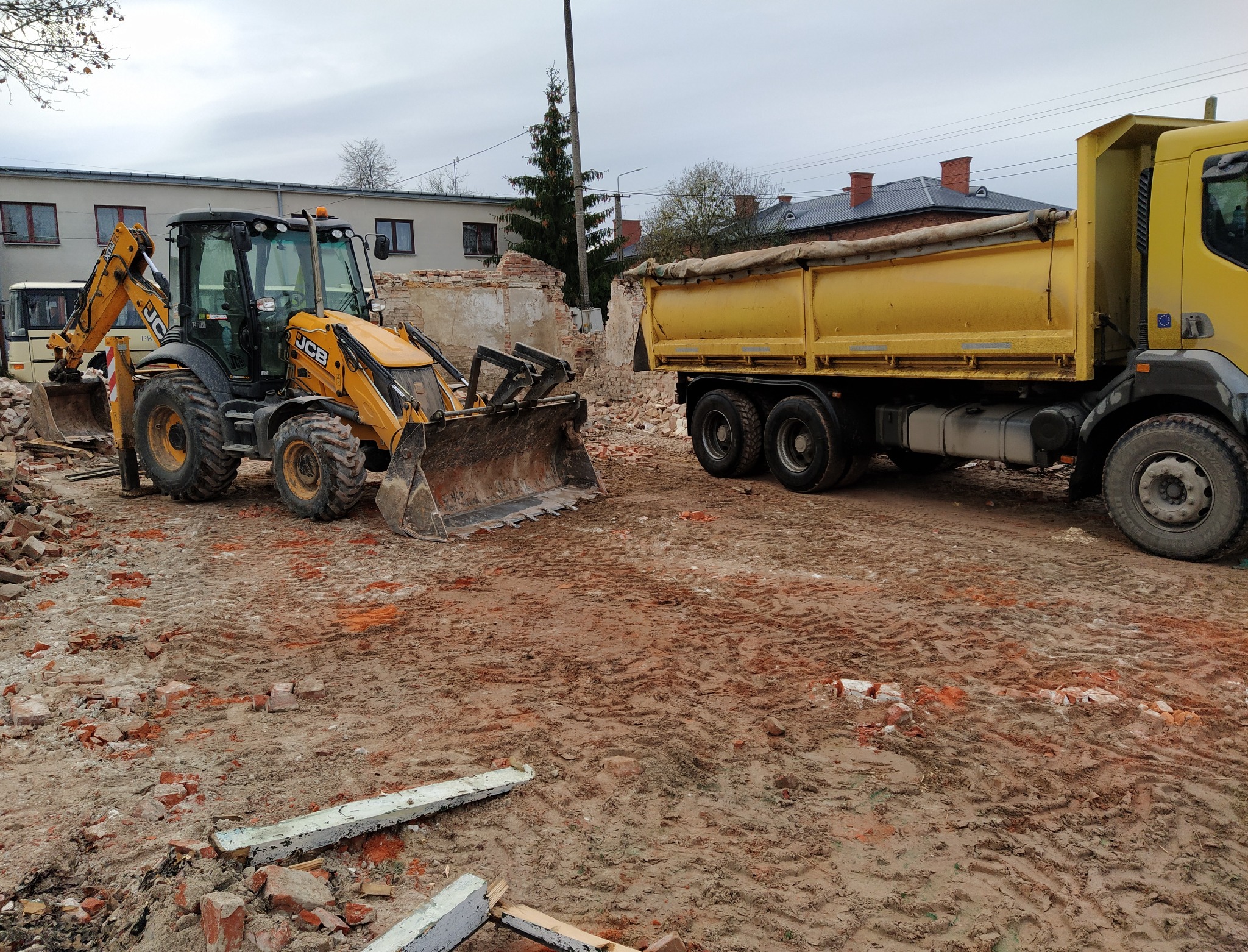 Żółta koparko-ładowarka JCB i wywrotka na placu budowy z gruzem i błotem, pozostałości po wyburzonej ścianie w tle.