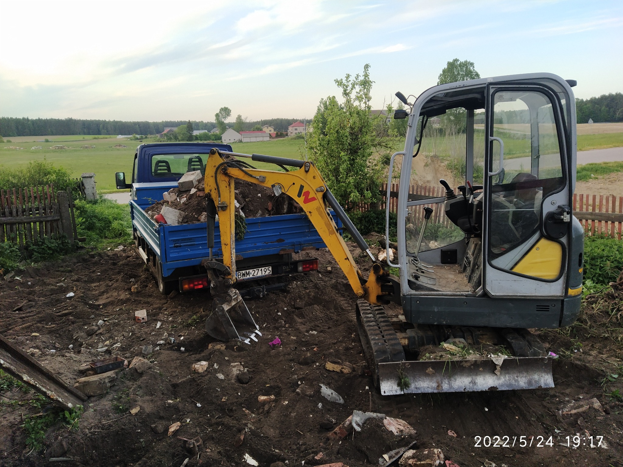 Mała koparka załadowuje gruz do niebieskiej ciężarówki na tle wiejskiego krajobrazu.