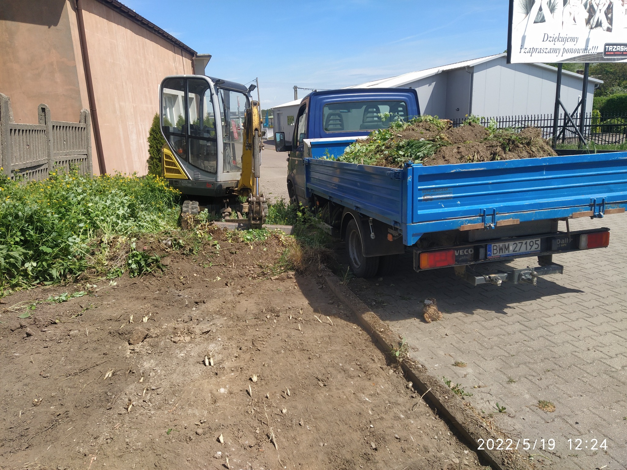 Niebieska ciężarówka z ładunkiem ziemi i zieleni obok małej koparki na placu budowy, widoczne ślady pracy w ziemi.