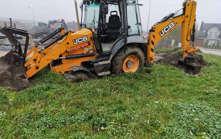 Żółto-szara koparko-ładowarka JCB w trakcie prac ziemnych na trawiastym terenie, widoczne ślady błota na oponach i łyżce, w tle zabudowania.