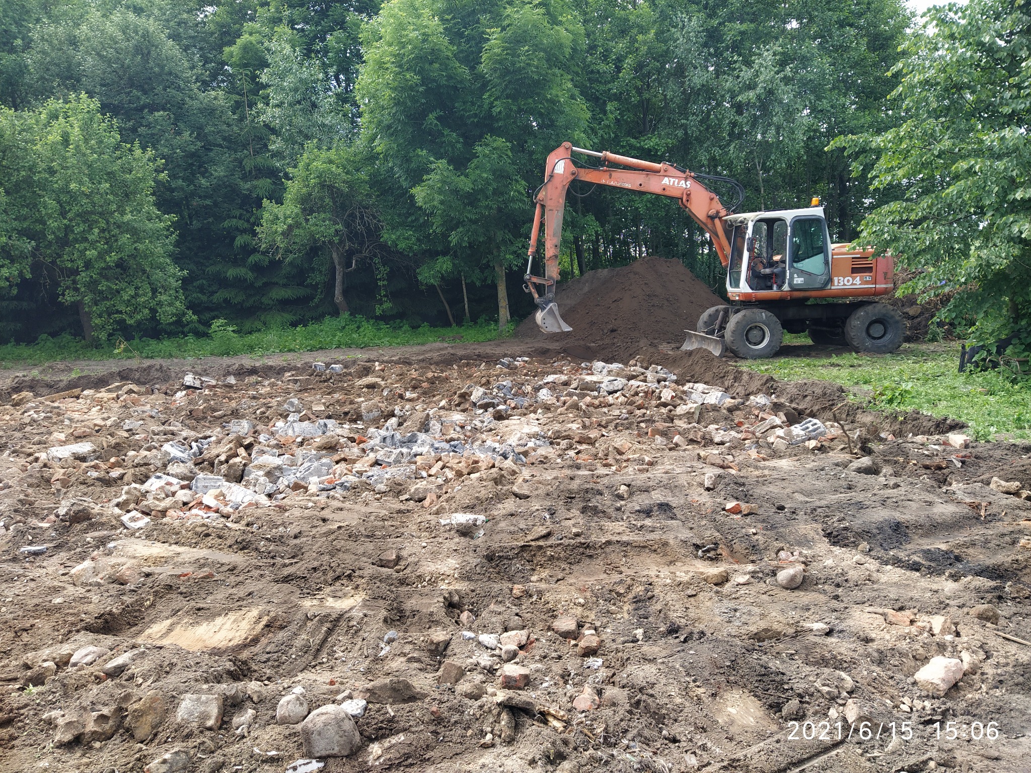 Pomarańczowa koparka Atlas 1304 LD na terenie budowy z gruzem i stertą ziemi na tle zielonego lasu.