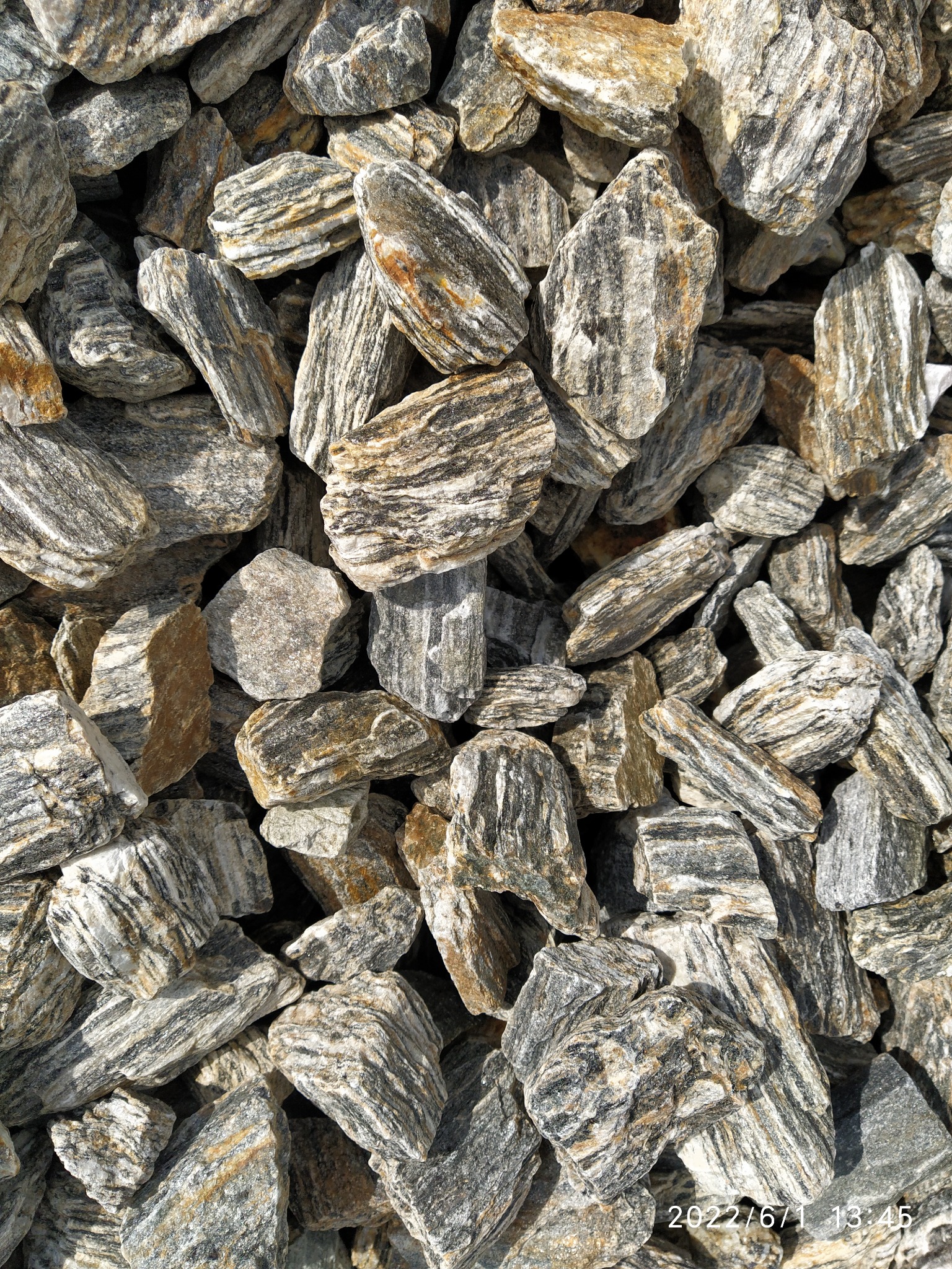 Kupa drobnych kamieni o nieregularnych kształtach i wyraźnych warstwach w odcieniach szarości, beżu i brązu, oświetlona naturalnym światłem słonecznym.