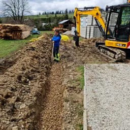 Wąski wykop wypełniony żwirem, obok żółta rura i sterta desek, w tle krajobraz z pagórkami, mężczyzna oparty o łopatę i żółta koparka JCB 55Z-1