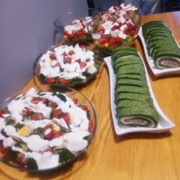 Szklane misy z sałatkami warzywnymi polanymi sosem i zielone roladki ze szpinakiem na białych prostokątnych półmiskach, ustawione na drewnianym stole.