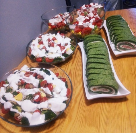 Szklane misy z sałatkami warzywnymi polanymi sosem i zielone roladki ze szpinakiem na białych prostokątnych półmiskach, ustawione na drewnianym stole.