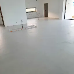 Jasnoszara, gładka podłoga w przestronnym, minimalistycznym wnętrzu w trakcie wykańczania, widoczne elementy instalacji podłogowej i surowe ściany.