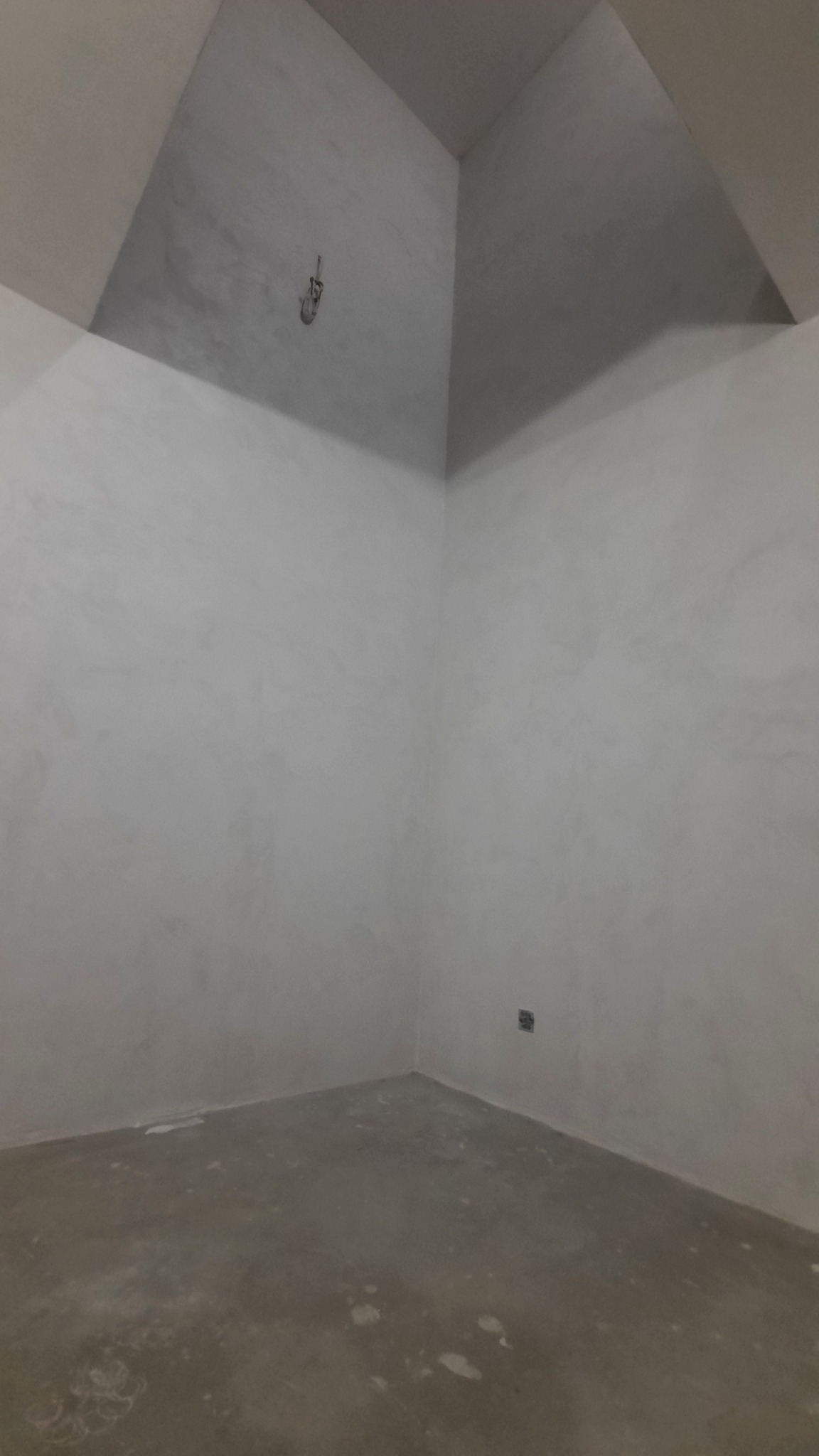Surowe wnętrze pomieszczenia w trakcie wykańczania, z szarymi ścianami i podłogą, widoczne ślady po pracach budowlanych i wystający przewód z sufitu.