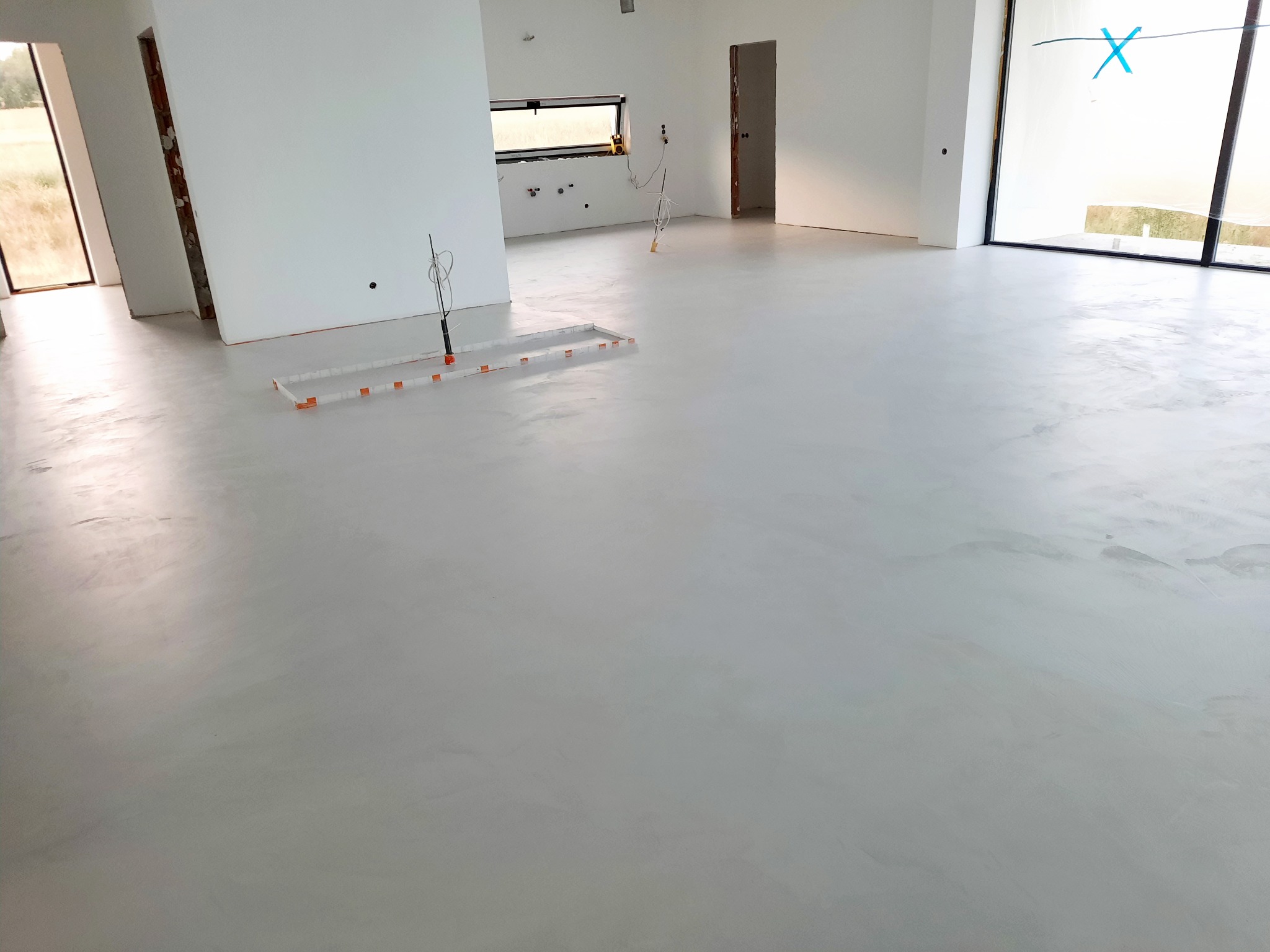 Jasnoszara, gładka podłoga w przestronnym, minimalistycznym wnętrzu w trakcie wykańczania, widoczne elementy instalacji podłogowej i surowe ściany.