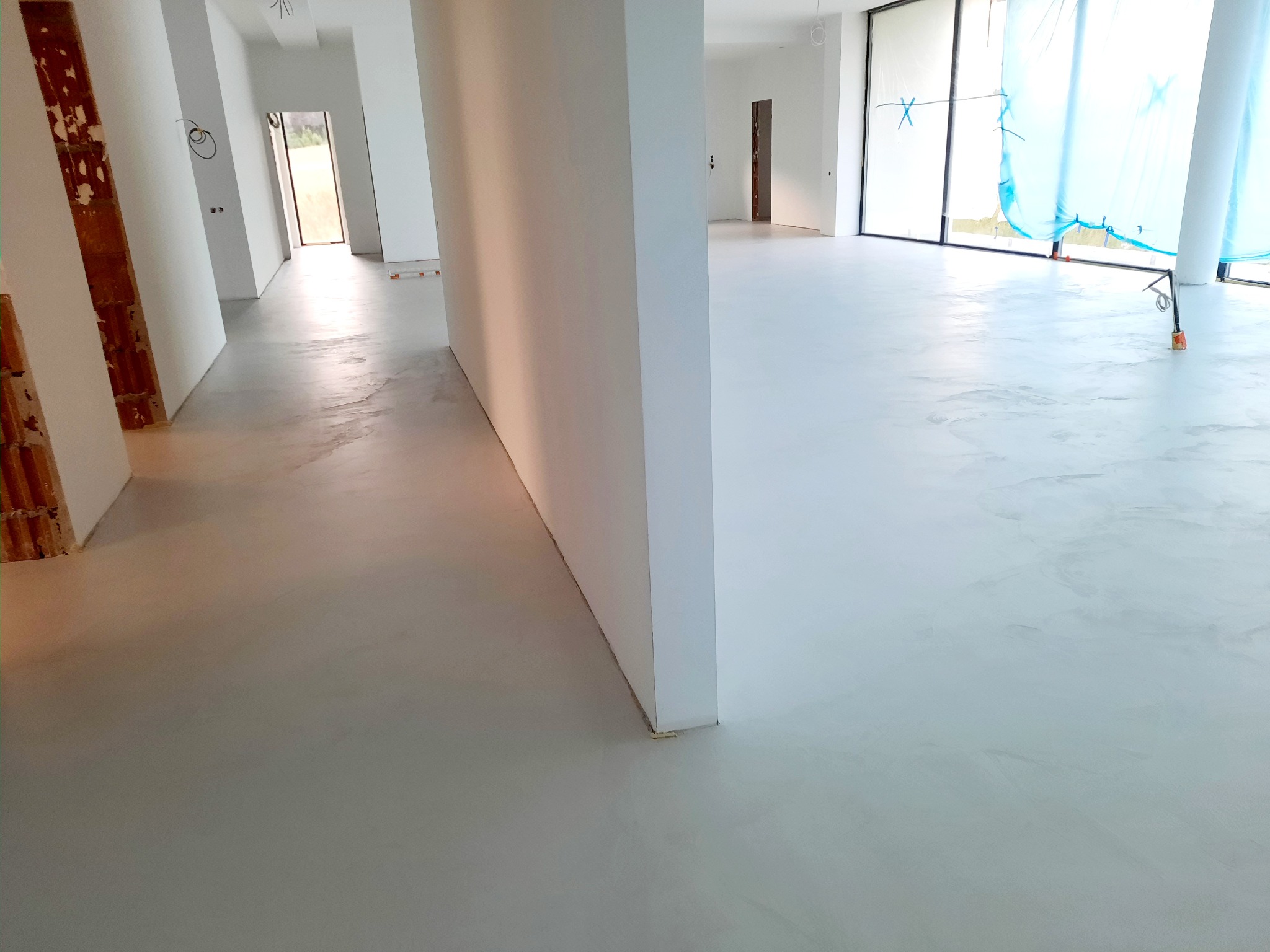 Jasna, gładka podłoga w przestronnym wnętrzu w trakcie wykańczania, widoczne surowe ściany i okna z folią ochronną.