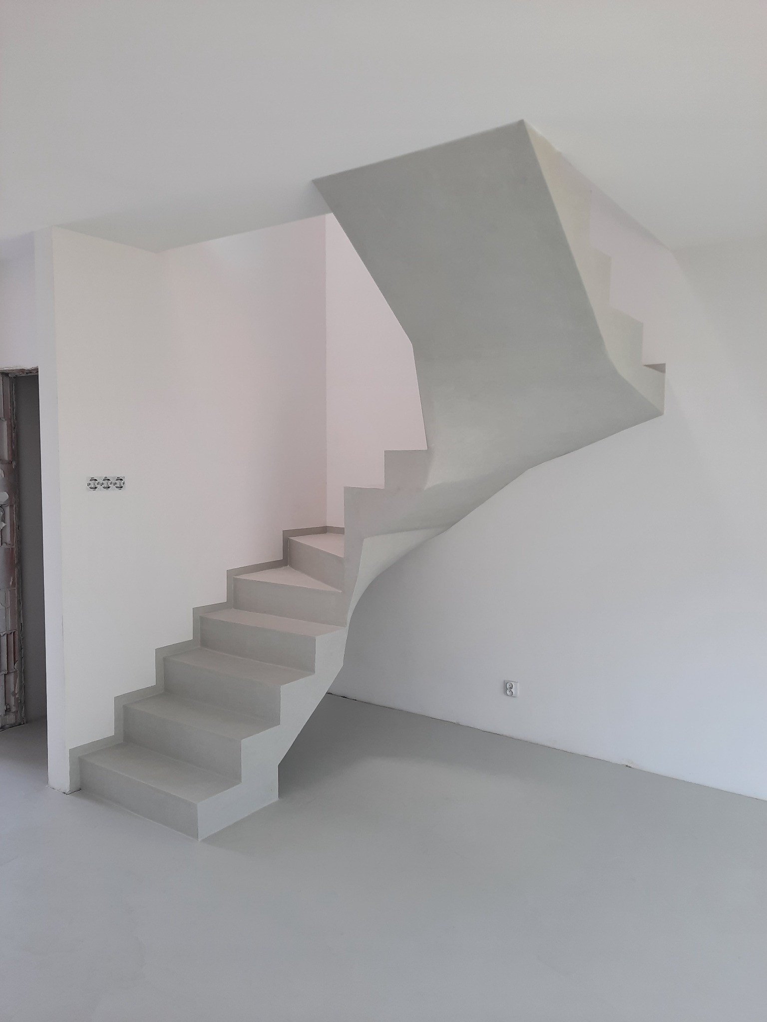 Minimalistyczne schody z mikrocementu w jasnym, surowym wnętrzu; widoczne gładkie powierzchnie ścian, podłogi i schodów w odcieniach szarości i bieli.