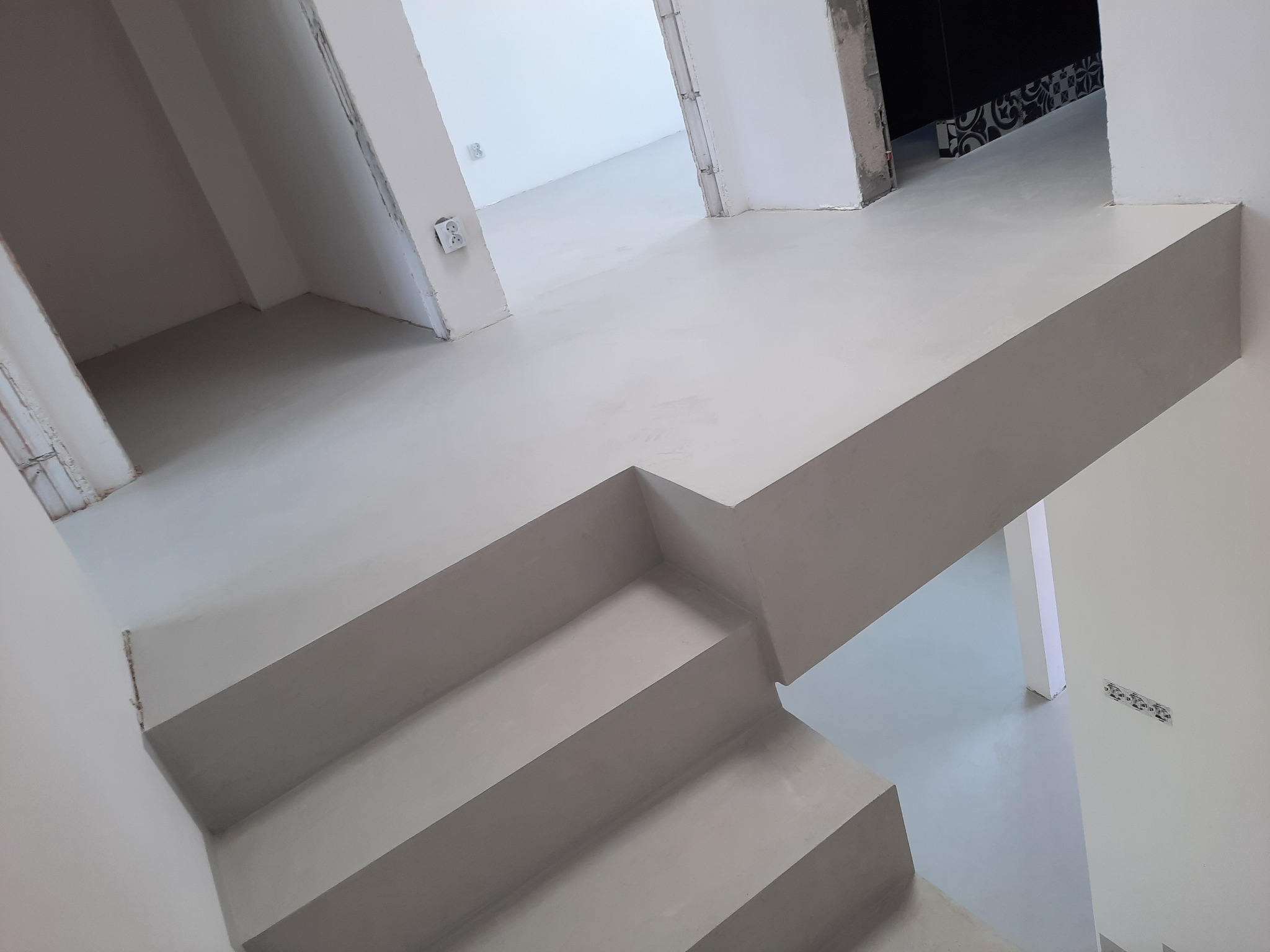 Jasnoszara, jednolita powierzchnia schodów i podłogi w surowym wnętrzu, widoczne gniazdka elektryczne i niedokończone wykończenia ścian.