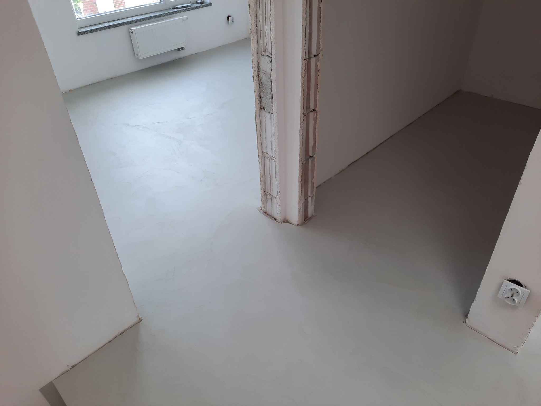 Świeżo wylana, jasnoszara posadzka w surowym wnętrzu, widoczny fragment kolumny konstrukcyjnej i gniazdko elektryczne przy podłodze.