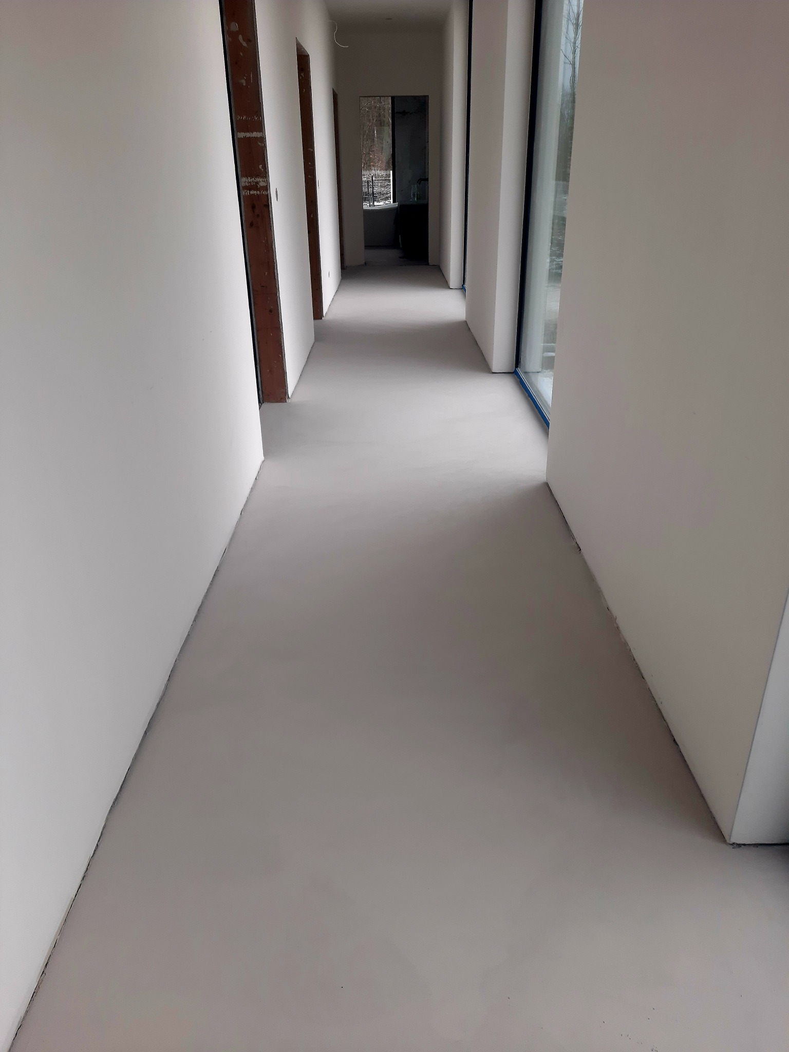 Długi korytarz z równą, szarą podłogą, ściany w kolorze złamanej bieli, widoczne surowe ościeżnice drzwiowe i duże okno po prawej stronie.