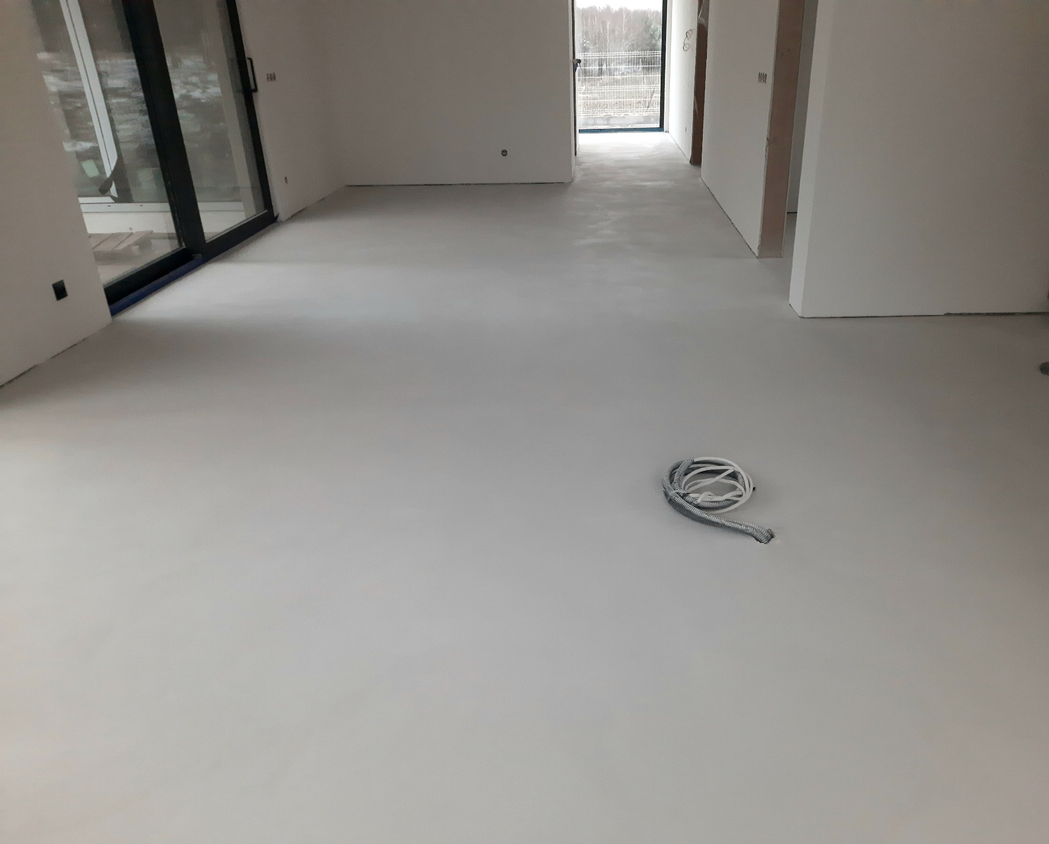 Jasnoszara, gładka podłoga w przestronnym, surowym wnętrzu z dużymi oknami; zwój kabli leży na posadzce w prawym dolnym rogu.