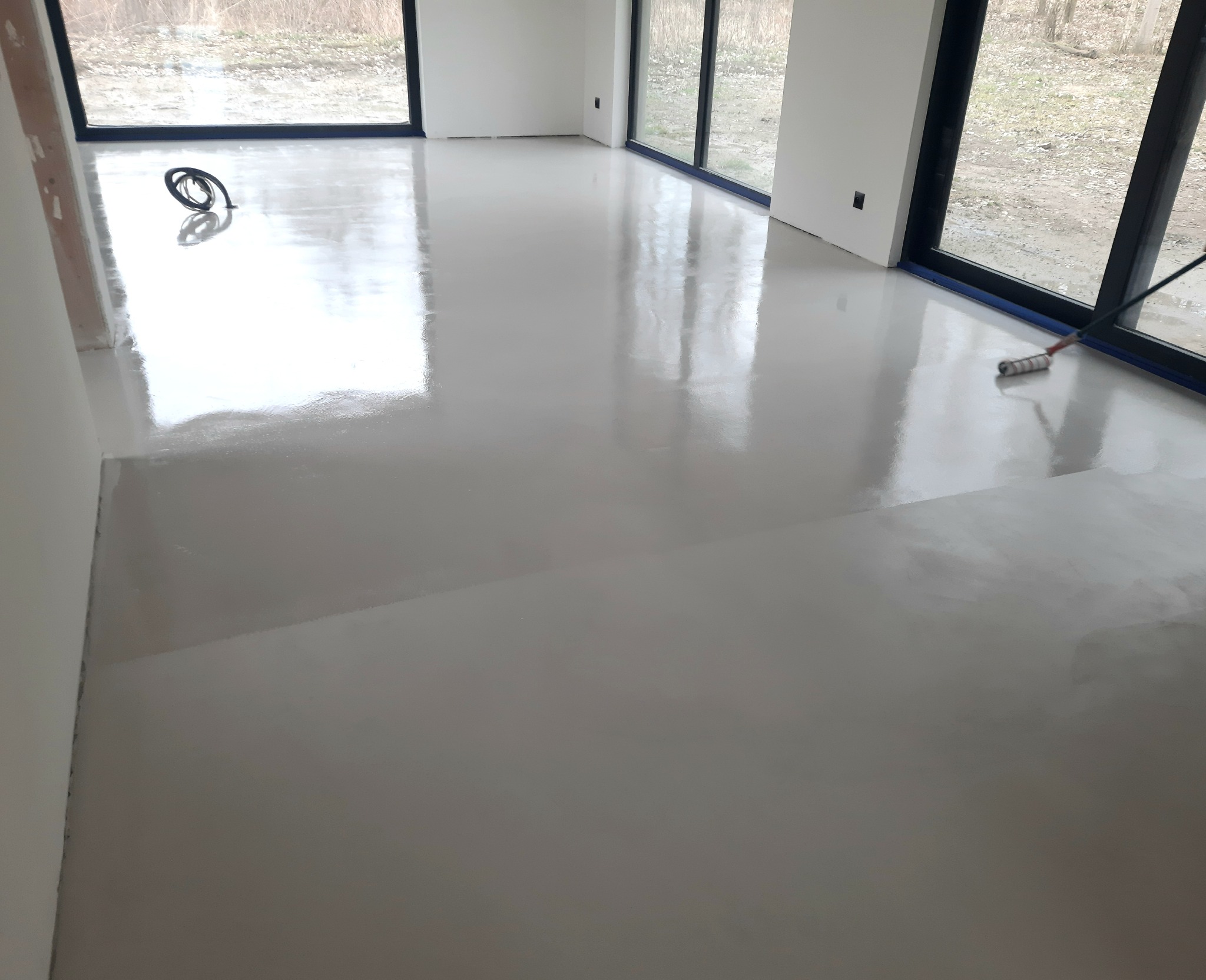 Jasnoszara, błyszcząca podłoga w trakcie aplikacji, widoczny wałek malarski przy oknie, kabel elektryczny na podłodze, duże okna z widokiem na zewnątrz.