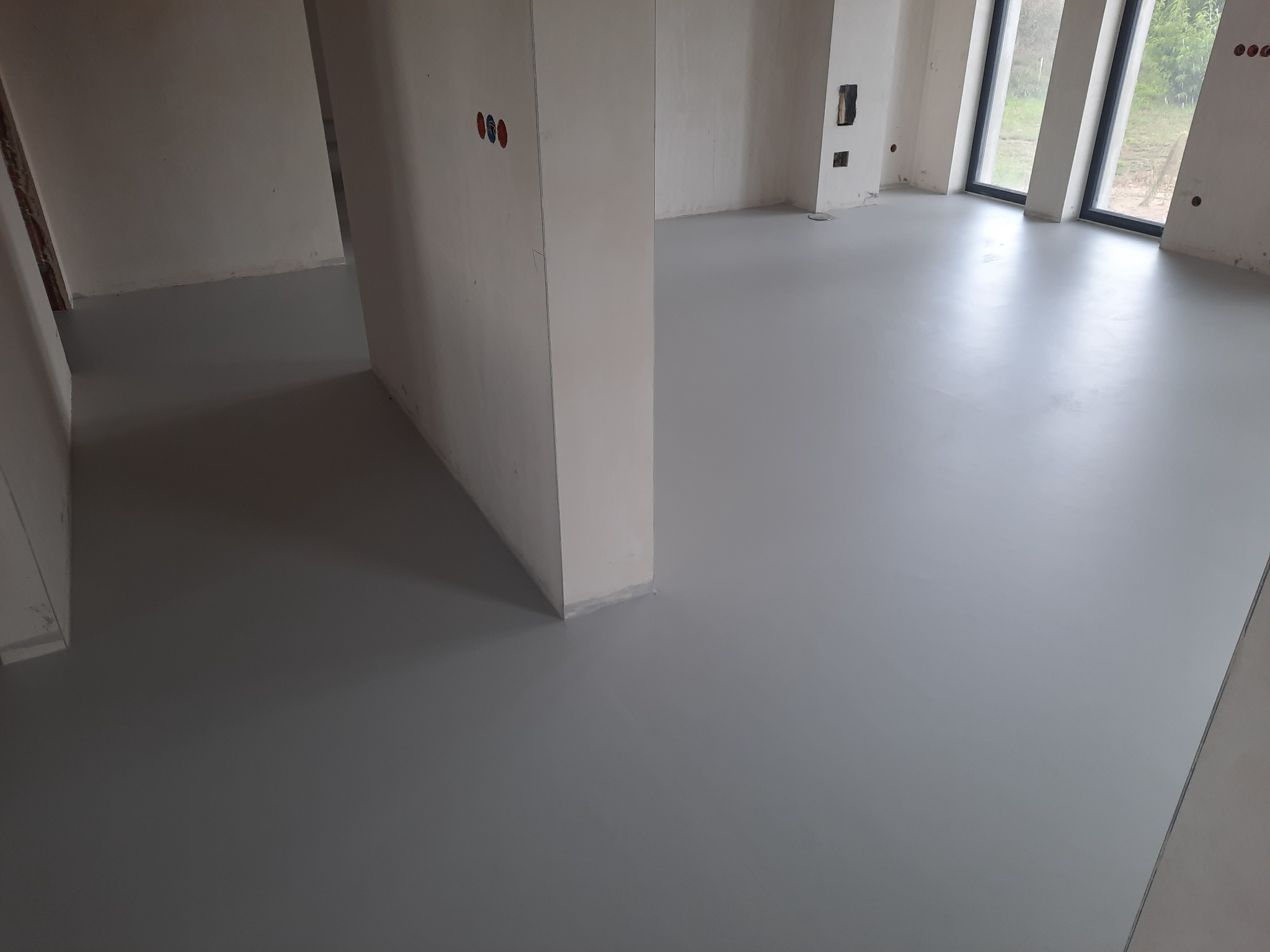 Szara, gładka posadzka żywiczna w surowym wnętrzu z widocznymi instalacjami elektrycznymi i dużymi oknami.