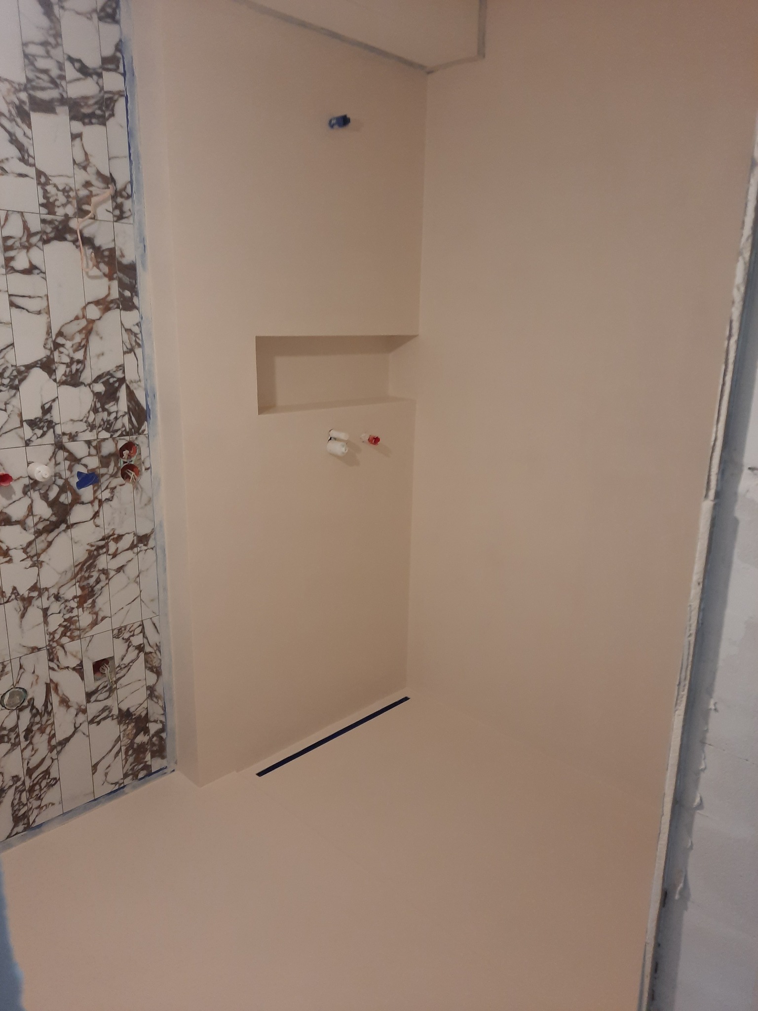 Przygotowana do użytku kabina prysznicowa z beżową podłogą i ścianami, z fragmentem ściany wyłożonej marmurowymi płytkami, widoczne przyłącza wodne i odpływ liniowy w podłodze.