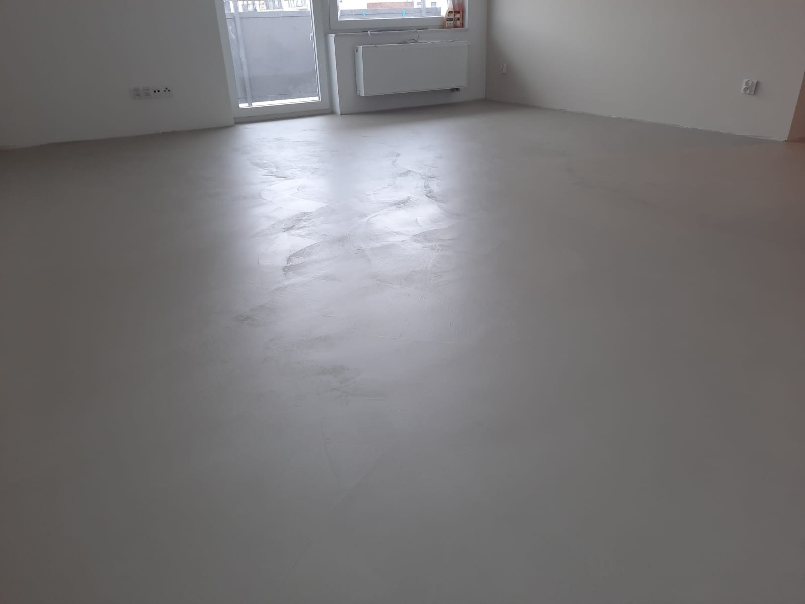 Jasnoszara, gładka posadzka z delikatnym efektem chmurkowania w pustym pomieszczeniu z oknem balkonowym i grzejnikiem.