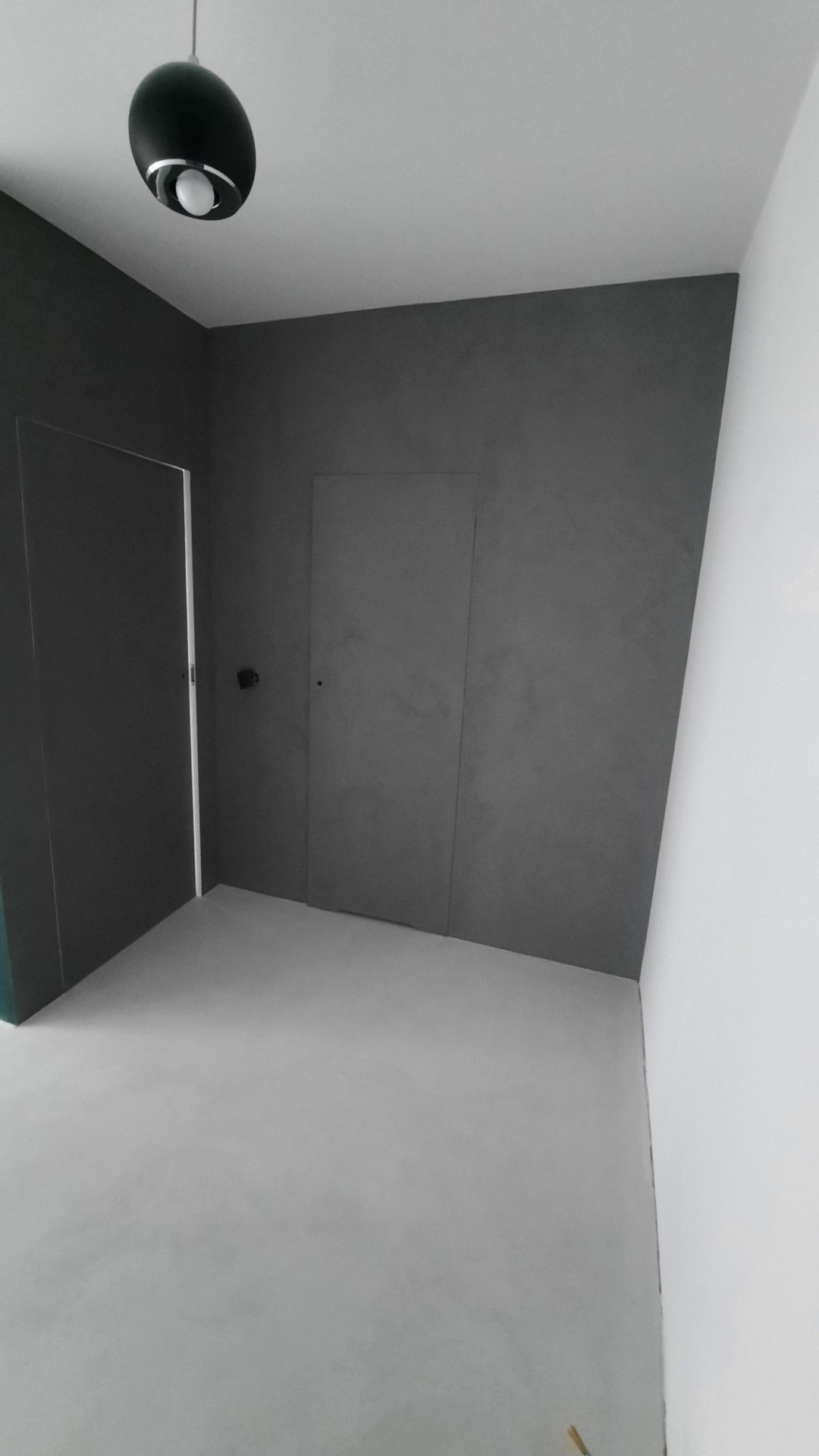 Wnętrze z szarą ścianą z ukrytymi drzwiami i jasną podłogą, oświetlone czarną, kulistą lampą sufitową.
