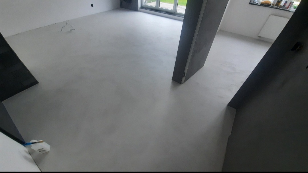 Jasnoszara, jednolita podłoga w nowym pomieszczeniu z dużym oknem i widocznym narzędziem malarskim w rogu.