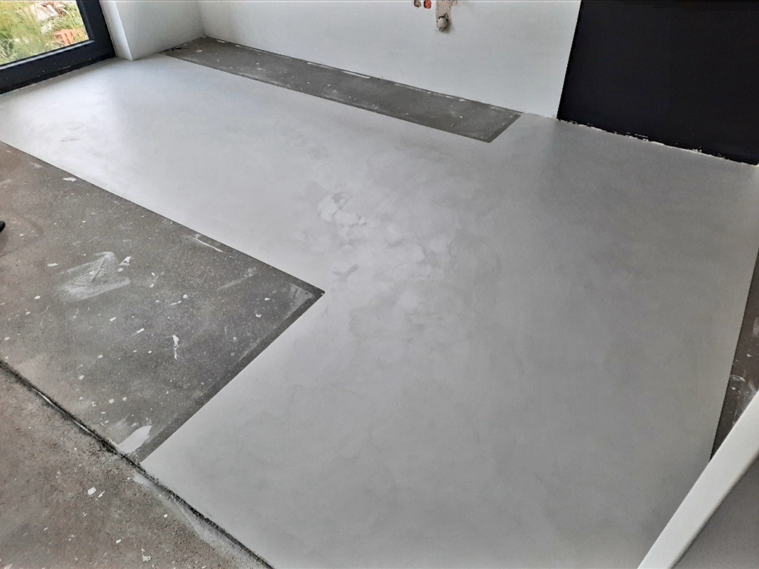 Szara, gładka podłoga w trakcie wykonywania, z widocznymi fragmentami surowego podłoża i śladami białej farby, w minimalistycznym wnętrzu z czarnym fragmentem ściany.