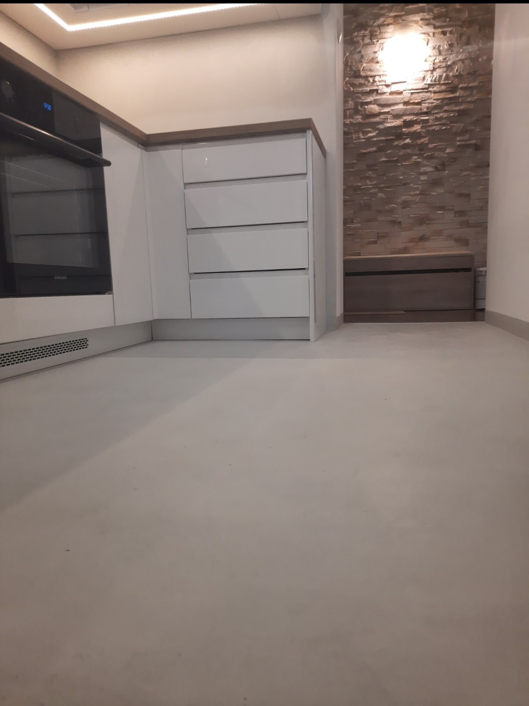 Jasnoszara, jednolita posadzka w kuchni z białymi szafkami i kamienną ścianą w tle, widoczna dolna część piekarnika i listwa przypodłogowa.