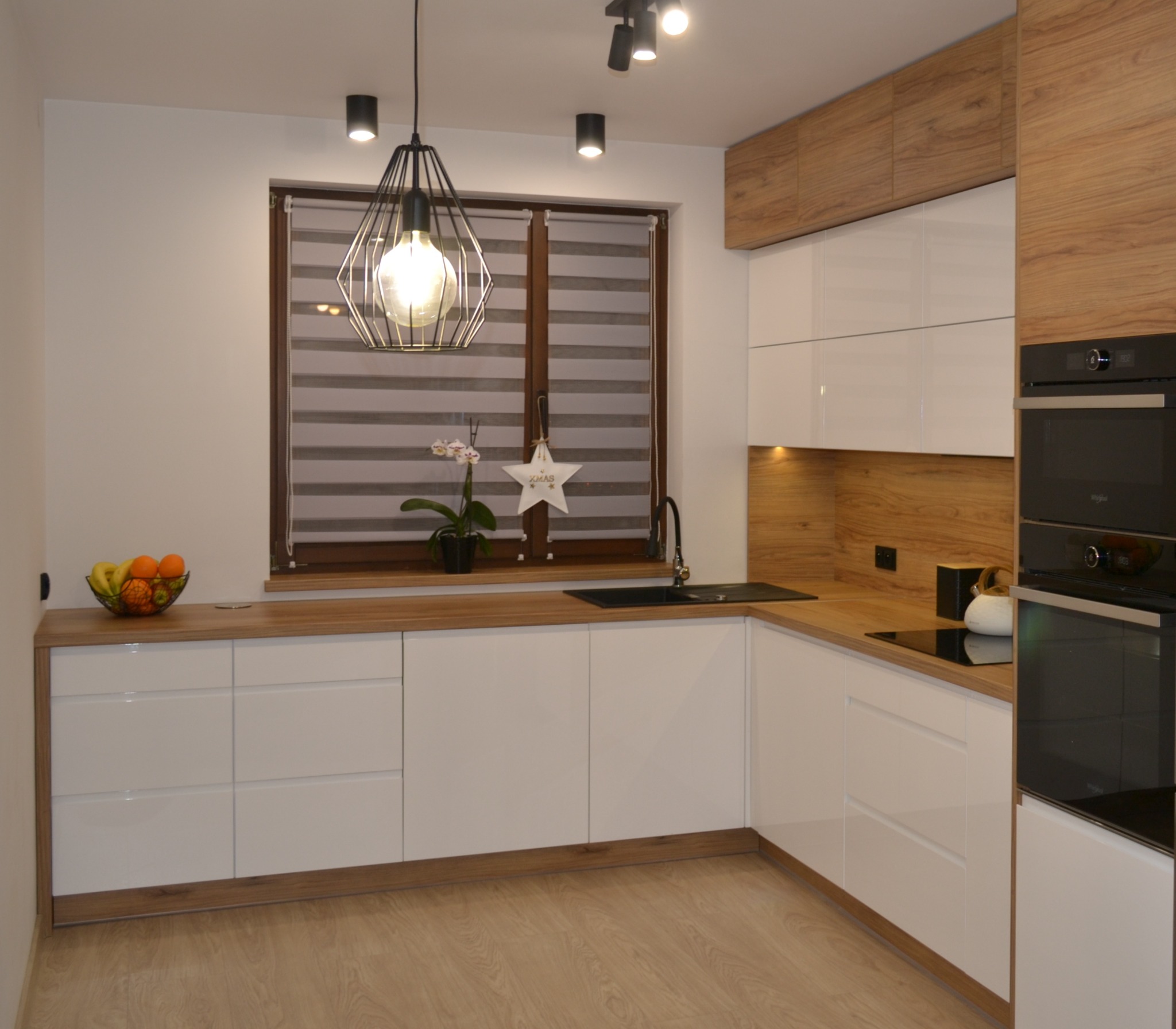 Nowoczesna, jasna kuchnia z białymi frontami i drewnianymi blatami, oświetlona lampą wiszącą z metalowym kloszem i halogenami sufitowymi, z widocznym czarnym zlewem i elementami dekoracyjnymi...