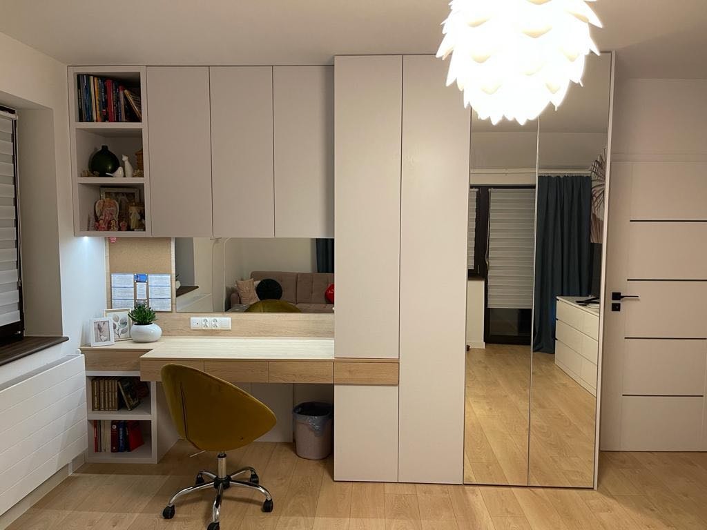 Białe biurko z drewnianym blatem i szafkami wiszącymi, zintegrowane z szafą z lustrzanymi drzwiami, oświetlone lampą w kształcie kwiatu, z żółtym krzesłem biurowym na kółkach.