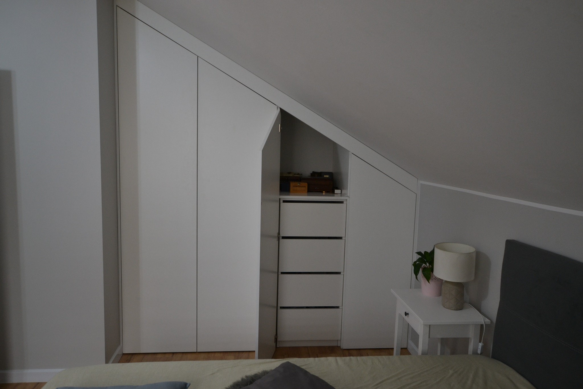 Biała szafa wnękowa z otwartymi drzwiami, dopasowana do skosu poddasza, widoczna z perspektywy łóżka w sypialni.