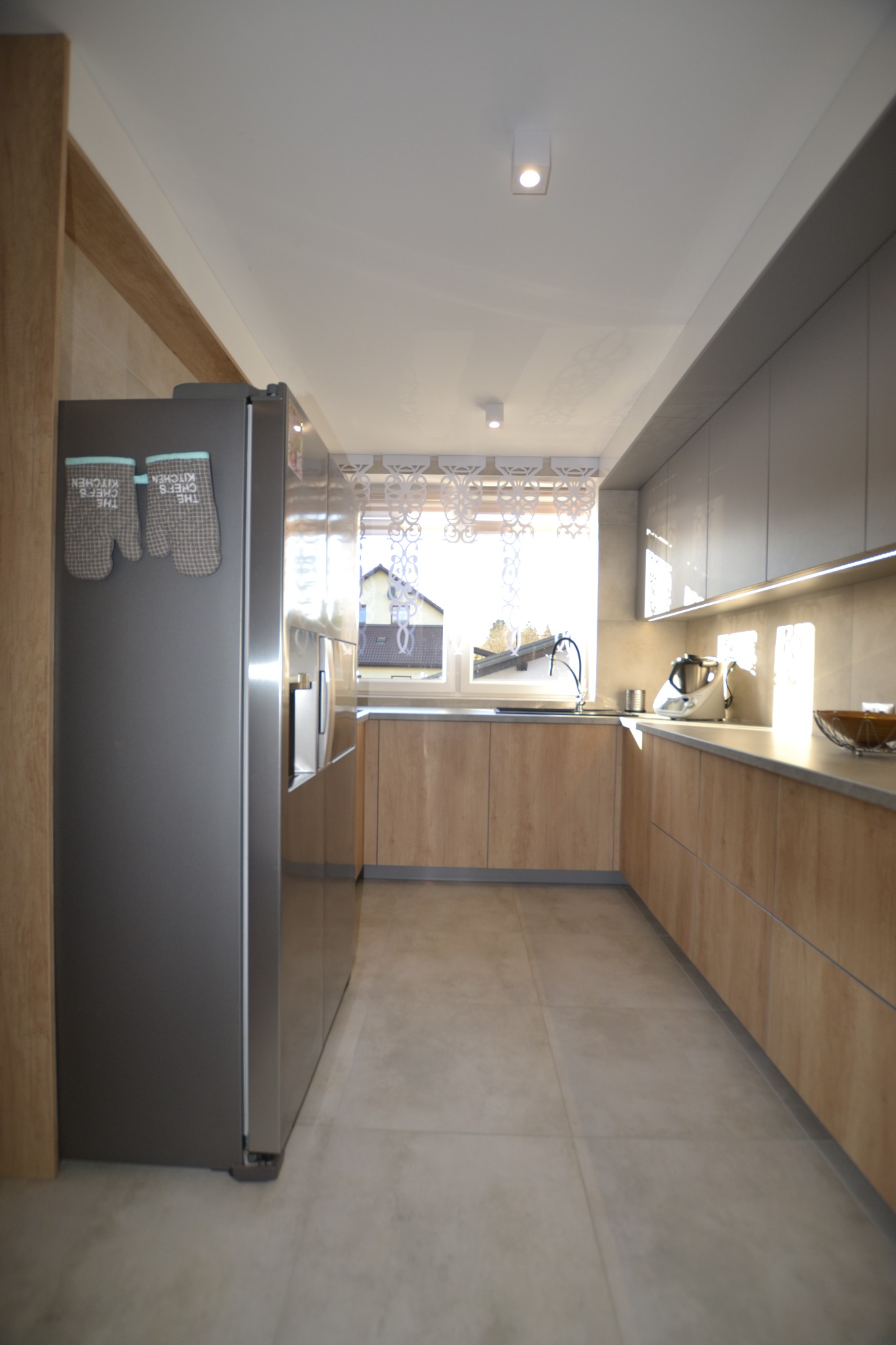 Wnętrze nowoczesnej kuchni z zabudową w odcieniach drewna i szarości, widok na okno z dekoracyjną firanką, na lodówce wiszą kuchenne rękawice.