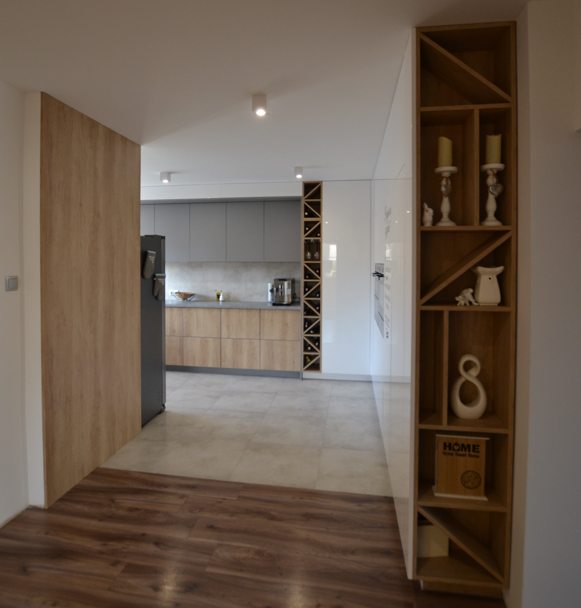 Nowoczesna kuchnia z szarymi szafkami górnymi i drewnianymi dolnymi, widoczna zza drewnianej ściany z wbudowanym regałem na wino i dekoracje.