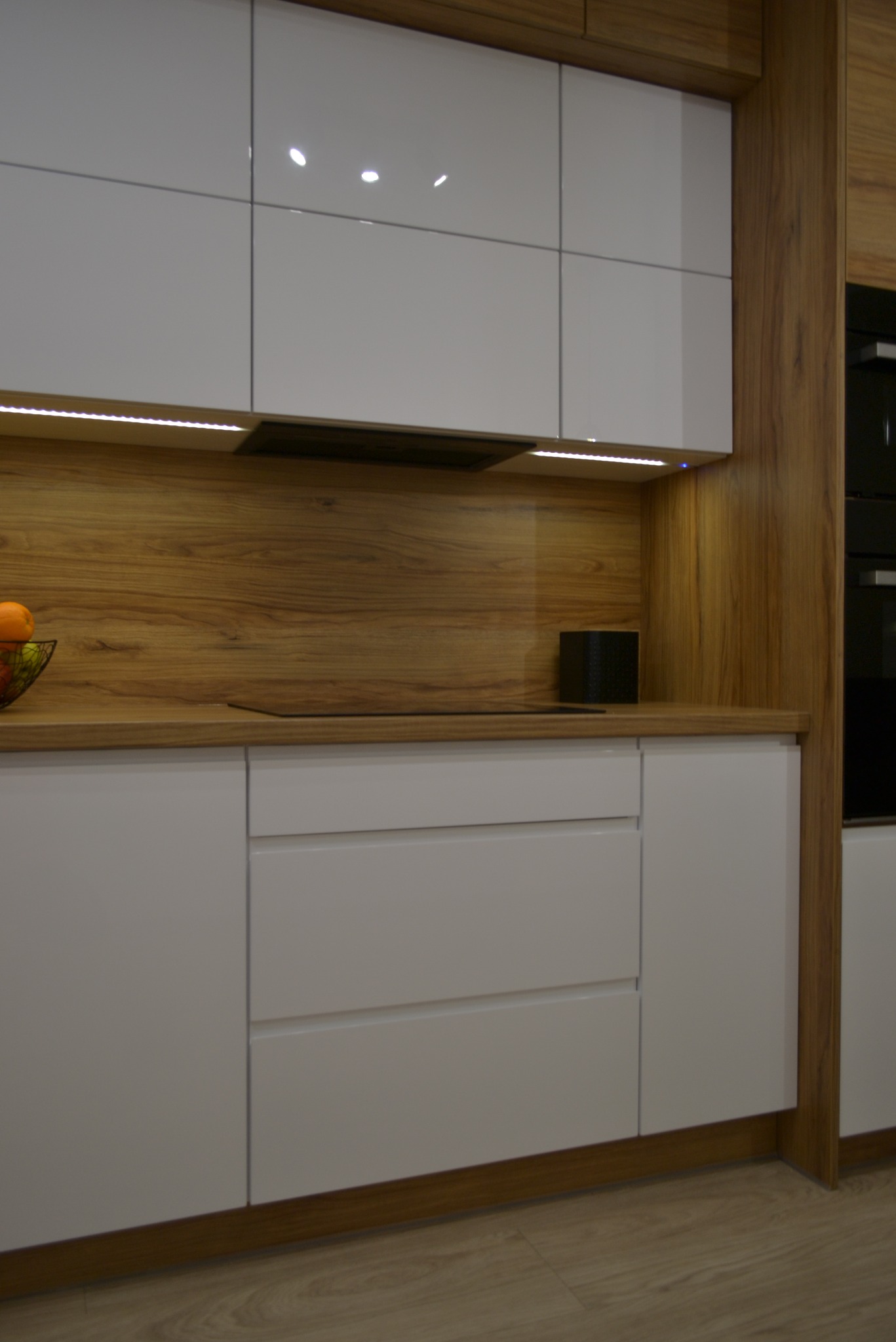 Nowoczesna zabudowa kuchenna z białymi, lakierowanymi frontami i drewnianym blatem, oświetlenie LED podszafkowe, minimalistyczny design, widoczny czarny głośnik na blacie.
