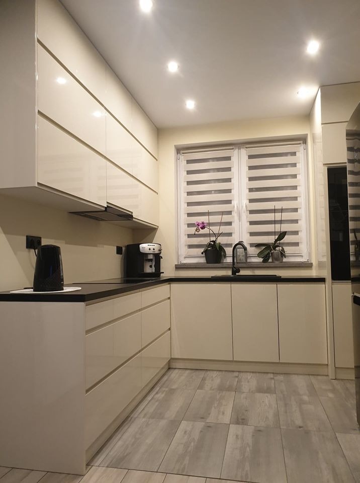 Nowoczesna kuchnia z kremowymi, lakierowanymi frontami, czarnym blatem, drewnopodobną podłogą i oknem z żaluzjami typu dzień-noc, na parapecie dwie doniczki z roślinami.