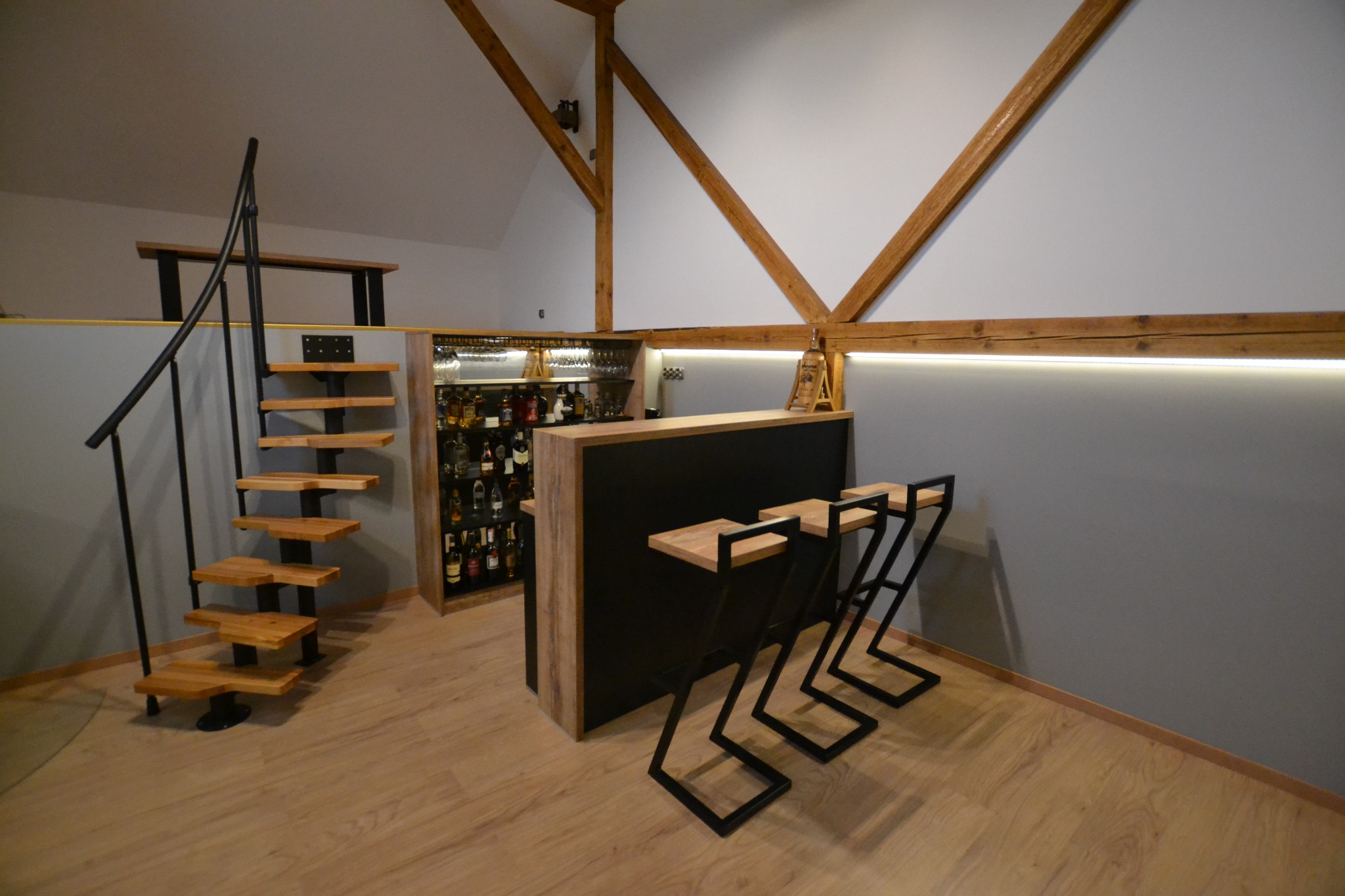 Nowoczesny bar z drewnianymi schodami prowadzącymi na antresolę, podświetlany barek z alkoholem, designerskie krzesła barowe na drewnianej podłodze w przestronnym wnętrzu z widoczną drewnianą...