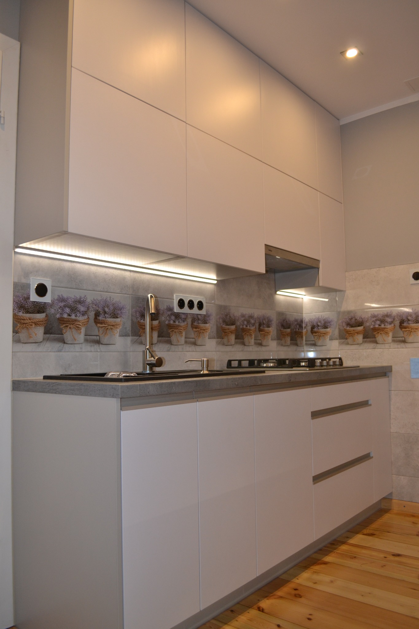 Jasna zabudowa kuchenna z gładkimi frontami, szarym blatem i płytkami z motywem lawendy nad blatem, oświetlenie LED pod szafkami wiszącymi i drewniana podłoga.