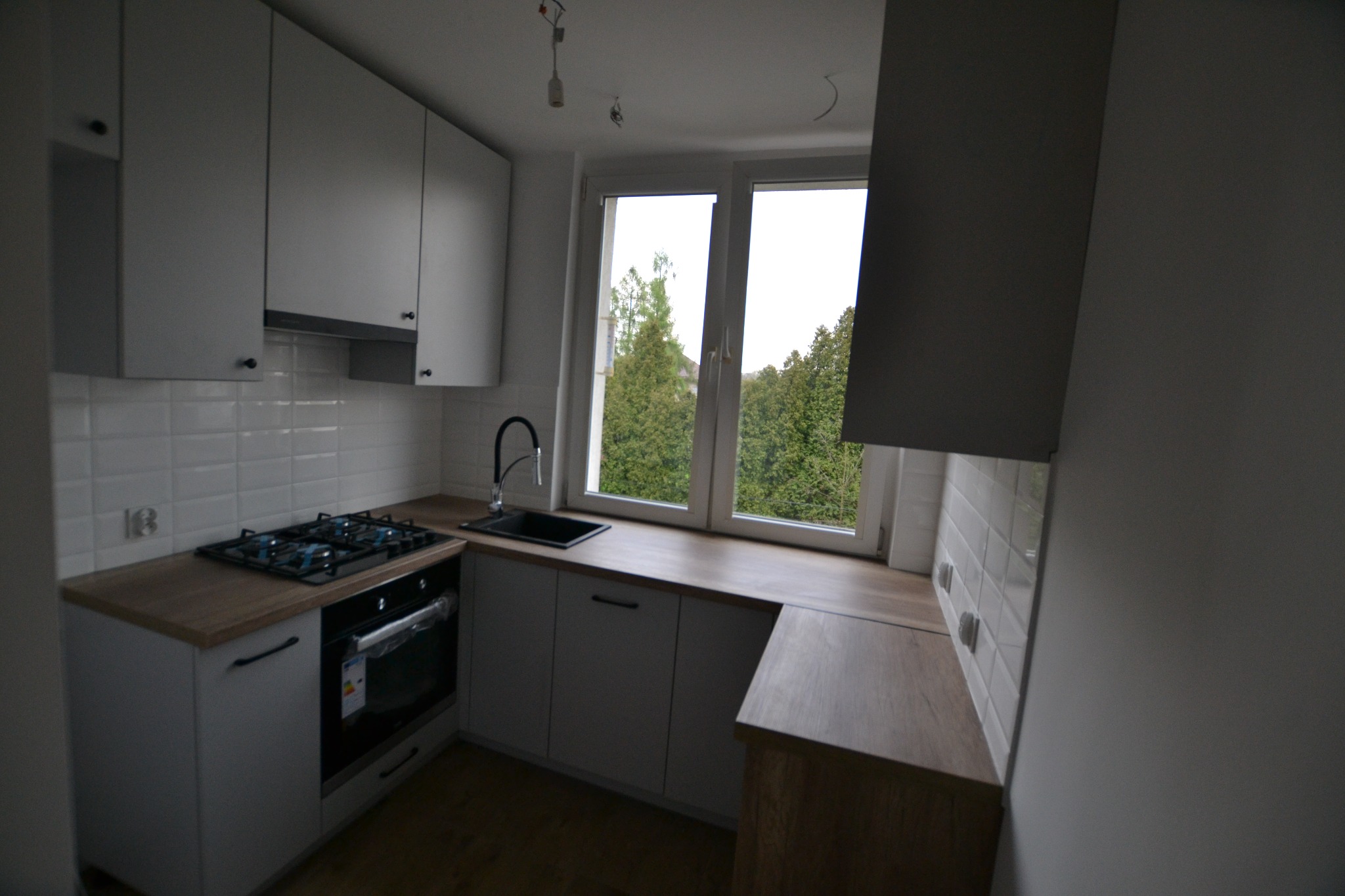 Nowoczesna kuchnia z białymi szafkami, drewnianym blatem i widokiem na zieleń za oknem. Płyta gazowa i piekarnik w zabudowie. Białe płytki na ścianie.