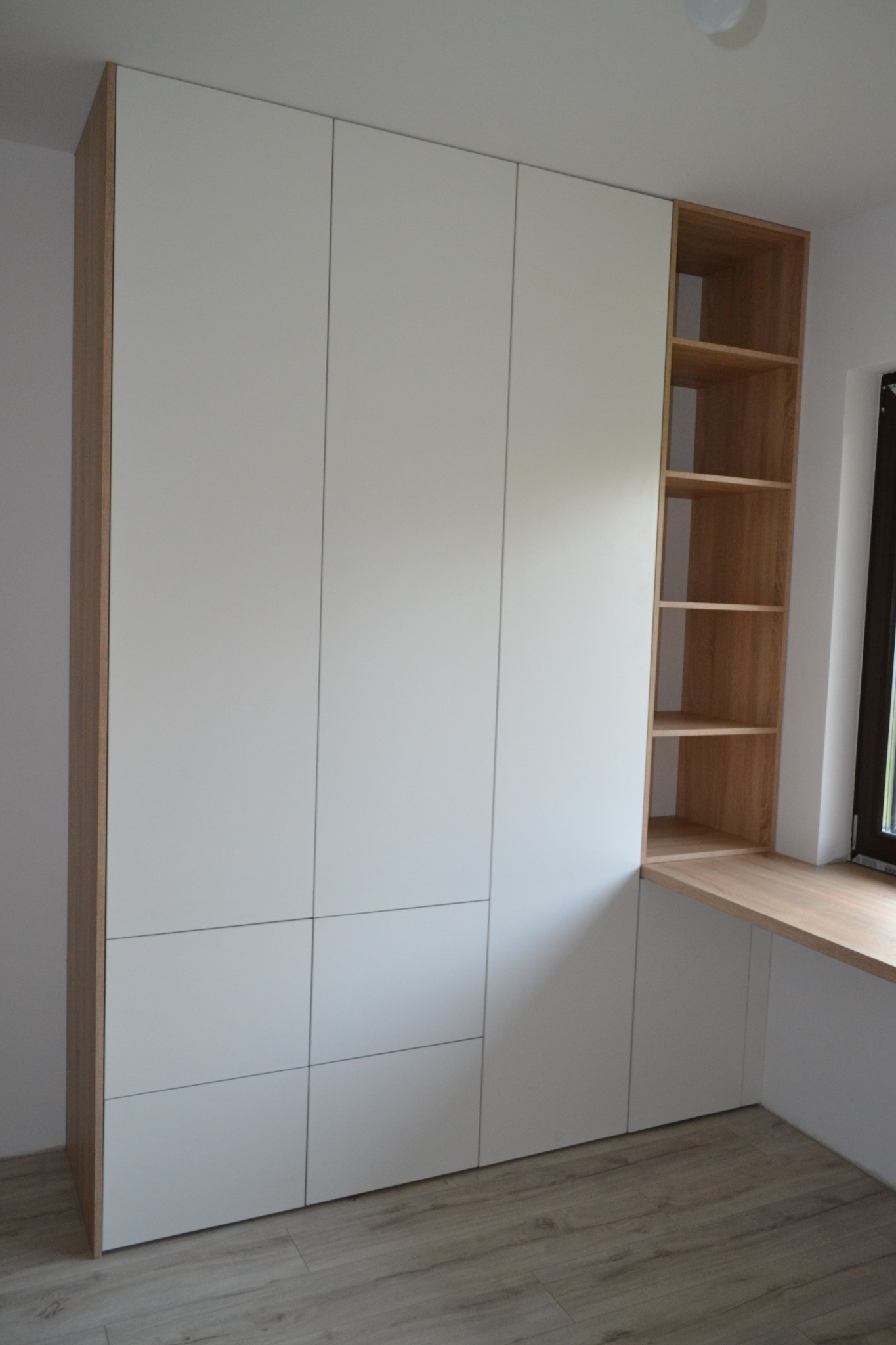 Biała szafa z drewnianymi elementami, wbudowana w ścianę obok okna, z drewnianymi półkami i blatem roboczym.