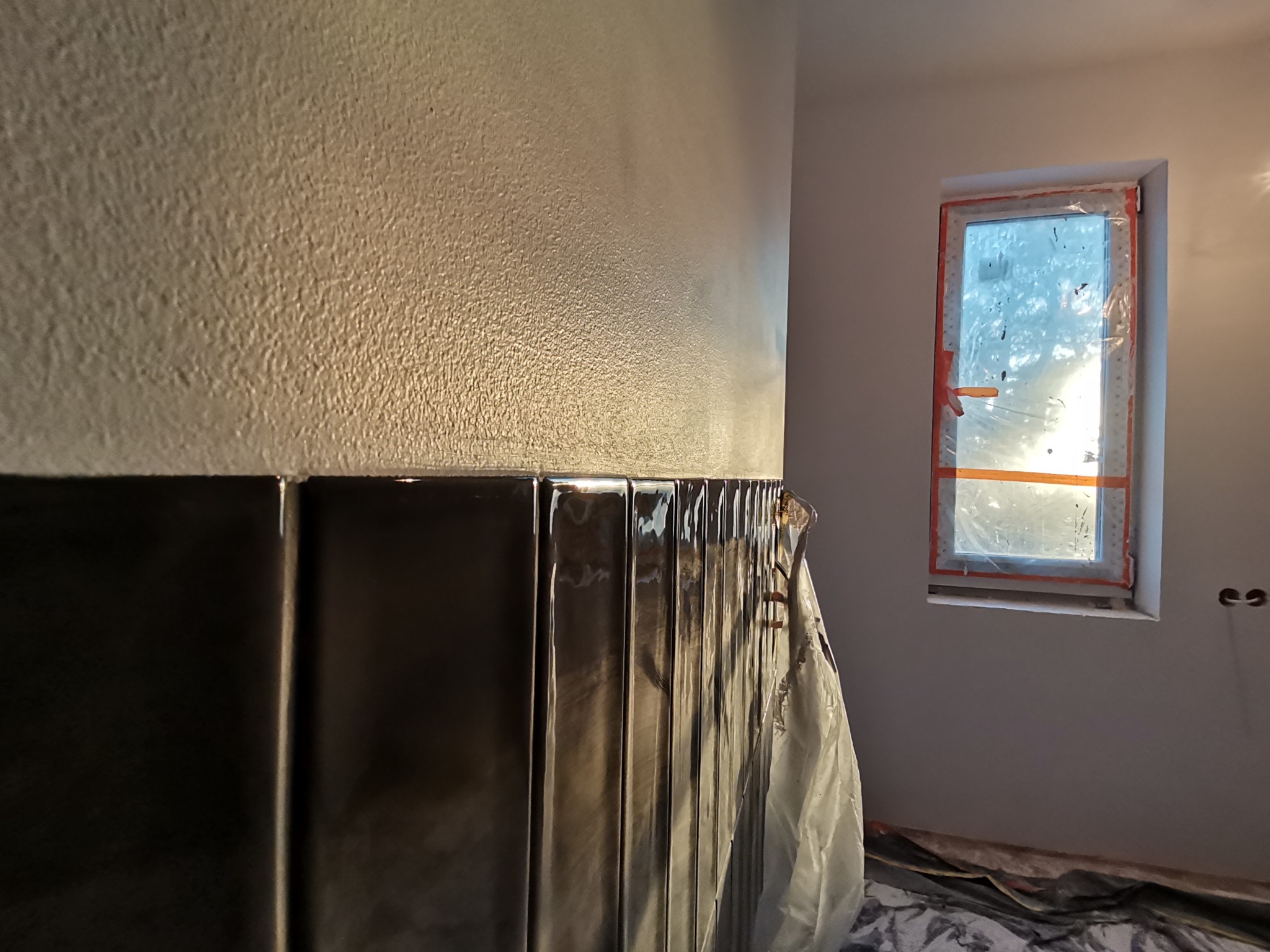 Świeżo pomalowana ściana z dekoracyjnymi, błyszczącymi panelami ułożonymi do połowy wysokości, zabezpieczone folią, w tle okno z pomarańczową ramą i zasłonięte folią, w pokoju w trakcie prac...